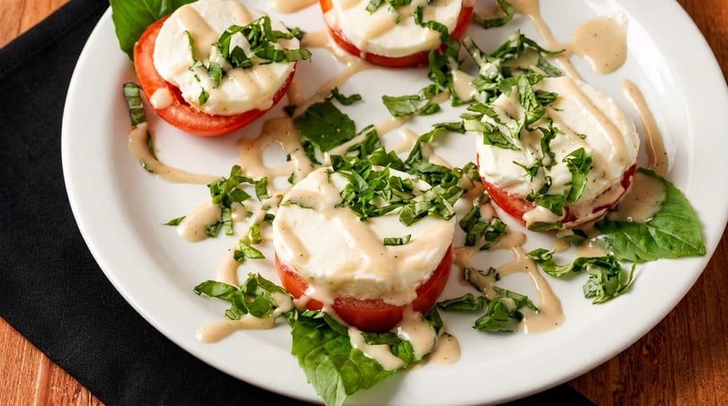 Caprese salad with sliced tomatoes, fresh mozzarella, basil, and a creamy dressing on a white plate.