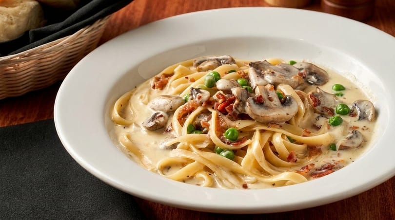 Creamy fettuccine pasta with mushrooms, peas, and bacon bits in a white bowl.